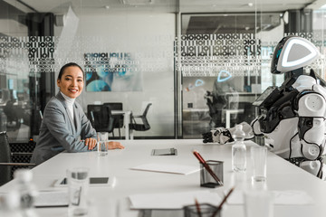 selective focus of smiling asian businesswoman looking at camera while sitting at desk near robot
