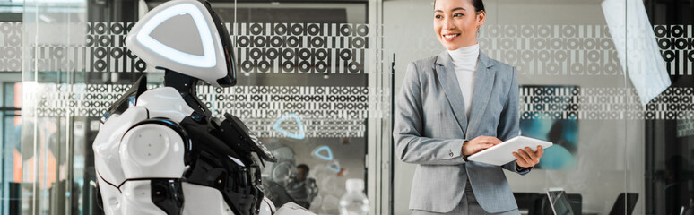 panoramic shot of smiling asian businesswoman holding digital tablet while looking at robot