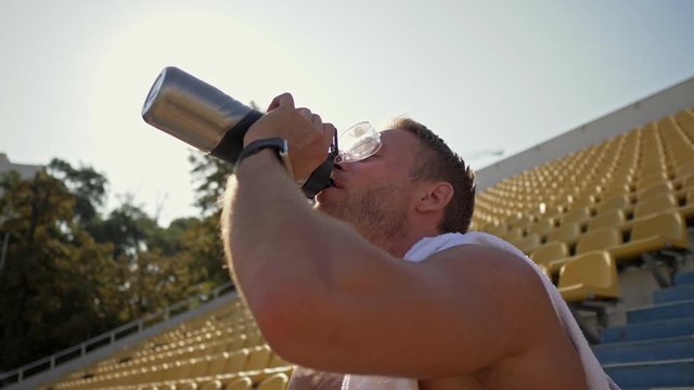 An attractive young shirtless sportsman with a towel on his shoulder is sitting and drinking water at the stadium looking aside