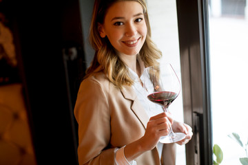 Single pretty young woman with glass of red wine standing near window and looking aside