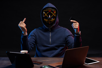 dangerous anonymous hacker man in scary mask sit at table with laptop, aggressive hacker in the hood of pullover show middle finger at camera isolated over dark space