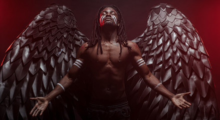 dark african angel with big black wings isolated, young serious muscular man wearing big wings on...