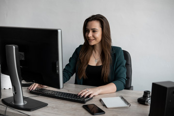 Brunette woman is working in front of a monitor in a office. Business woman working at computer at coworking space. Portrait of happy lady typing at workplace. Female professional working with pc.