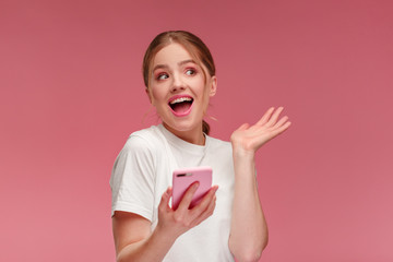 The best day ever. Surpised young redhead woman holding pink smartphone, smiling and expressing...