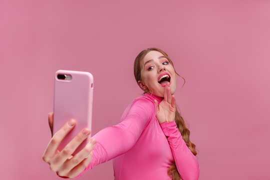 Funny young redhead woman making selfie. Smiling girl wearing pink blouse holding pink smartphone, making faces on camera, posing for selfie isolated on pink background.