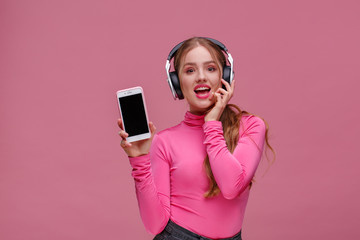 Funny redhead girl showing blank screen mobile phone on camera. Young smiling woman wearing earphones demonstrating smartphone display isolated over pink background. Copy space. App for your business