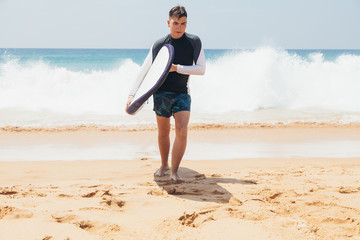 Fototapeta na wymiar Handsome teenage boy with surfboard coming out from the ocean at sandy beach on surf line.