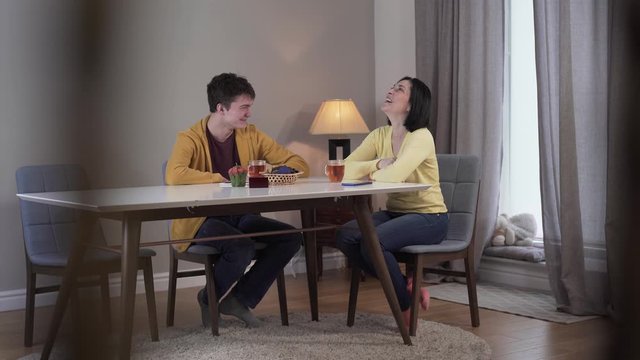 Shooting behind balusters of smiling Caucasian young boy and adult woman talking at the table and laughing. Joyful mother and teenage son chatting at home on weekends. Lifestyle, joy, communication.