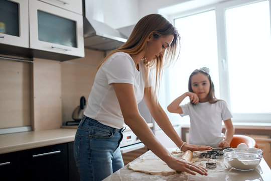 young fair-haired woman teaching her daughter to roll the dough. close up side view photo, kid wearing jeans and ehite T-shirt looking at the camera