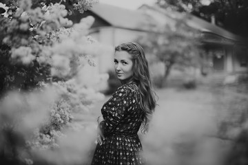 Beautiful redhead young woman posing in a long dress by a blooming lilac in the spring black and white colors