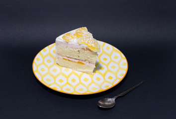 Piece of holiday cake with white cream and juicy apricot on white and yellow plate on black background