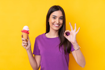 Young brunette woman with a cornet ice cream over isolated yellow background showing ok sign with fingers