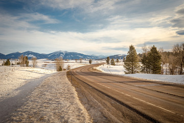 Empty highway with snowy fields on either side with vast mountain range in distance on beautiful day