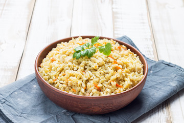 Vegetarian stewed side dish of brown rice with onions, carrots and celery greens in a bowl on a light wooden background. Traditional national diet cuisine. Copy space