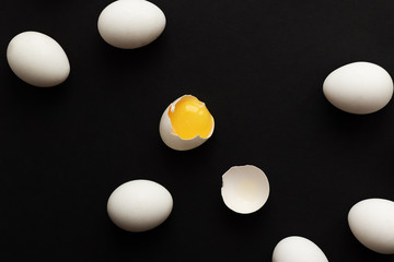White eggs lie scattered on a black background. One egg is broken and the yolk is visible. Minimalistic design and color trend 2020. Top view