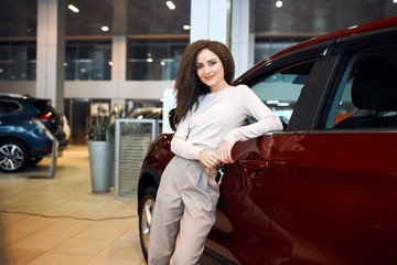 Obraz na płótnie Canvas Beautiful young business woman in stylish clothes is smiling and looking at camera while leaning on a red car in a motor show, close up photo, sale , discount, trade, industry