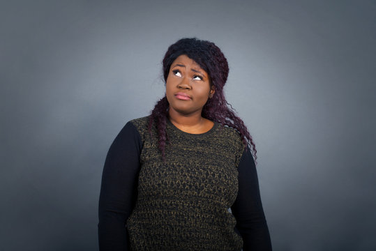 Troublesome puzzled Caucasian woman frowns face, bites lip, raises eyebrows, looks up, has problems, dressed in fashionable clothes, isolated over gray background, has regret look.