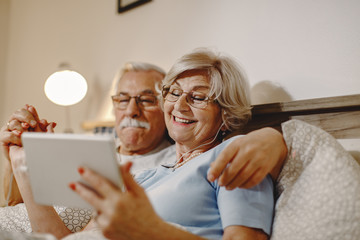Happy senior woman and her husband using digital tablet in bedroom.
