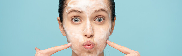 panoramic shot of funny woman with cleansing foam on face pointing on cheeks, isolated on blue
