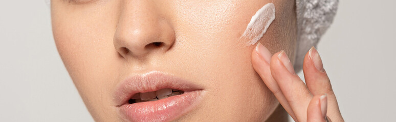 panoramic shot of young woman applying cosmetic cream on face, isolated on grey