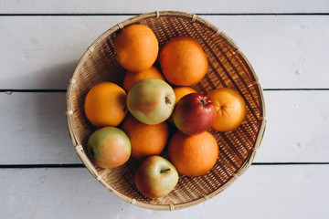  fresh fruits (apples and oranges) in a straw plate on a white wooden background close-up