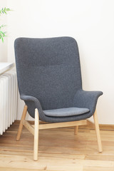 .gray armchair with wooden legs on a wooden floor near a heating battery..