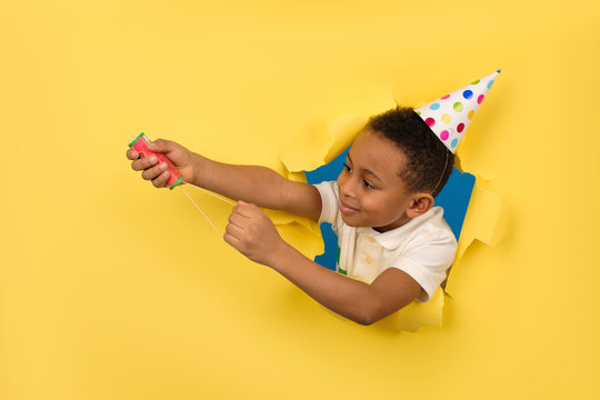 Happy African American Boy In White Polo Shirt And Holiday Hat Blows Up A Clapperboard In His Hands On A Yellow Studio Background With Torn Paper Edges. Birthday Or New Years Eve, Celebration Concept.