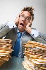 Overwhelmed office worker holding his head in shock at the large piles of paperwork on his desk