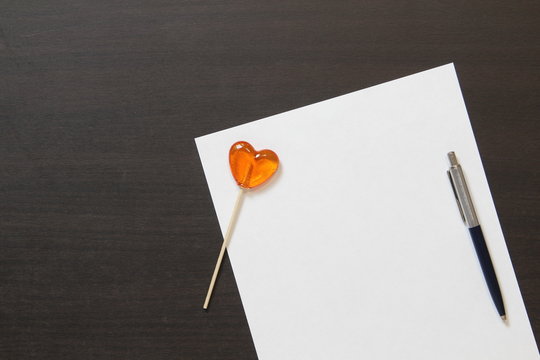 Template of white paper with pen and heart shaped caramel on dark wenge color wooden background.