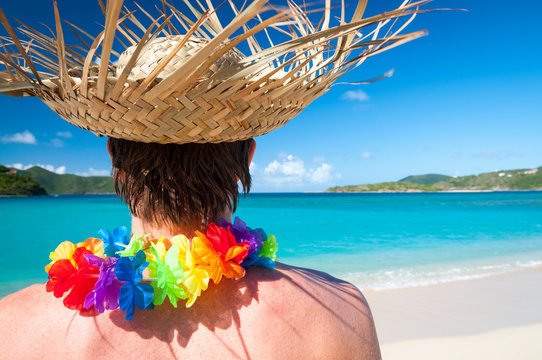 Unrecognizable tourist wearing woven palm frond hat and rainbow flower lei standing in front of a vibrant blue tropical paradise 