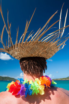 Unrecognizable tourist wearing woven palm frond hat and rainbow flower lei standing in front of a vibrant blue tropical paradise 