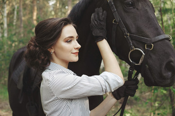 Beautiful girl jockey rider with a black horse, dressed in a light shirt in a green forest Park. The concept of horse riding.
