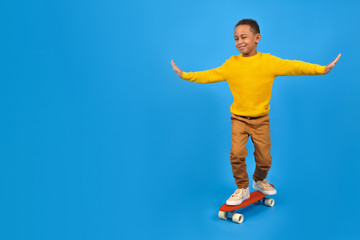 Fototapeta na wymiar Happy boy is African American riding a red skateboard balancing his arms placed in sides for a balance-clad yellow sweater and brown pants against a blue studio background. free space for text