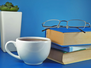 Stack of books with glasses on top and tea cup and succulent in white flowerpot from behind on blue background. Learning and education concept.              