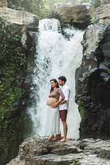 Fototapeta na wymiar Childbirth in Bali. Young pregnant couple travel on Tegenungan waterfall. Family harmony values. Happy together, pregnancy travel lifestyle. Beautiful morning nature. Maternity concept.
