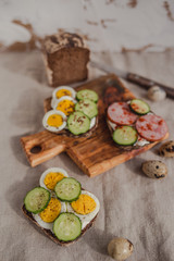 Set of sandwiches with rye bread, egg, ham, cucumber, cream cheese, seeds and spices on a wooden board. Rustic breakfast.