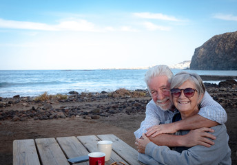 A happy senior couple embracing each other on the beach. Sitting at a wooden table with two cups of coffee. Two real people
