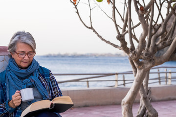 Fototapeta na wymiar Portrait of a senior woman enjoying her free time by reading a book sitting near the beach and holding a cup of tea. One relaxed people. Horizon over sea