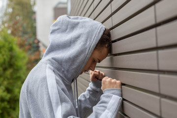 worried, sad guy teenager in a sweatshirt with a hood stands near the wall and beats his fist, the concept of teenage problems and psychology