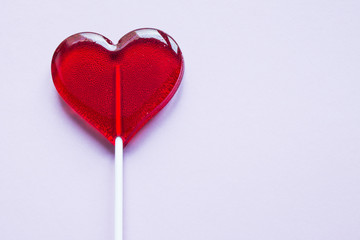 Sweet transparent Lollipop in the shape of a red heart on a pale pink background with copy space