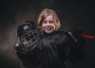 Young blonde sporty boy, ice hockey player, posing in a dark studio for a photoshoot, wearing an ice-skating uniform while holding his helmet, hockey stick and smiling on camera
