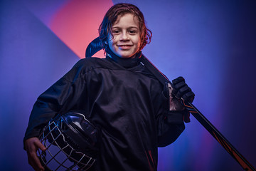 Young blonde fit boy, ice hockey player, posing in a bright neon studio for a photoshoot, wearing an ice-skating uniform while wearing his helmet, holding a hockey stick in his hand and looking
