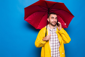 Young handsome bearded man in yellow raincoat with red umbrella talking by mobile phone isolated...