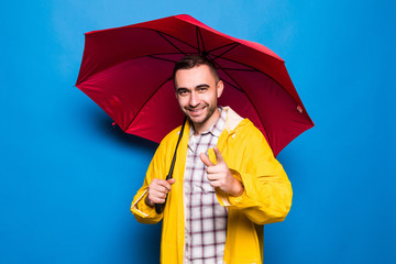 Young handsome bearded man in yellow raincoat with red umbrella pointed on you isolated over blue...