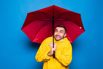 Young handsome bearded man in yellow raincoat with red umbrella cover from rain get cold isolated over blue background