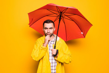 Young handsome bearded man in yellow raincoat with red umbrella thinking isolated over orange background