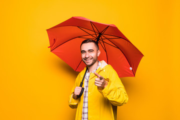 Young handsome bearded man in yellow raincoat with red umbrella pointed on you isolated over orange...