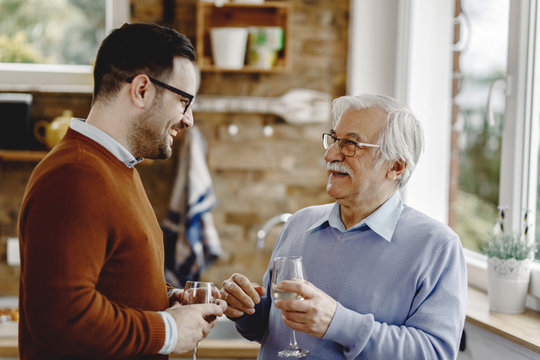 Happy Mature Man Talking With His Adult While Drinking Wine In The Kitchen.