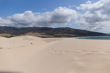 Fototapeta na wymiar landscape of a golden sand beach with dunes, in the distance you can see the mountains and the blue sea on a day with blue sky and white clouds