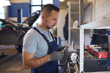 Fototapeta premium Side view portrait of bearded car mechanic choosing tools while repairing vehicle in garage shop, copy space